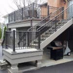 Image of a back porch with a concrete deck and a storage shed beneath with a two level staircase and wrought iron handrails.